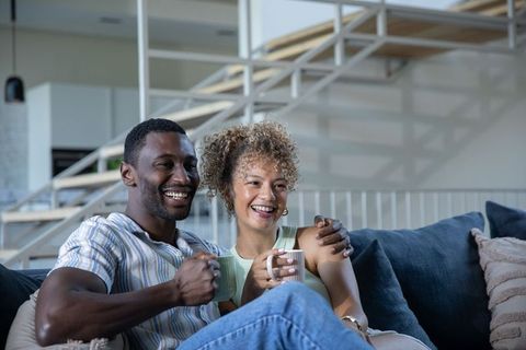 African american couple relaxing on dark blue sofa holding coffee mugs in modern living room