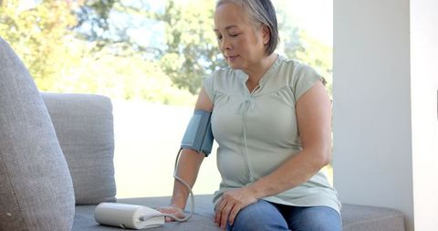 Image of a senior Asian woman calmly using a blood pressure monitor while seated on a comfortable couch. Perfect for illustrating healthcare articles, wellness programs focused on elderly patients, or home health monitoring systems for seniors.