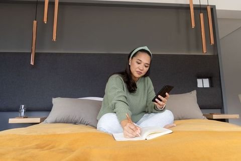 Asian woman multitasking on bed using smartphone while writing notebook in cozy modern bedroom