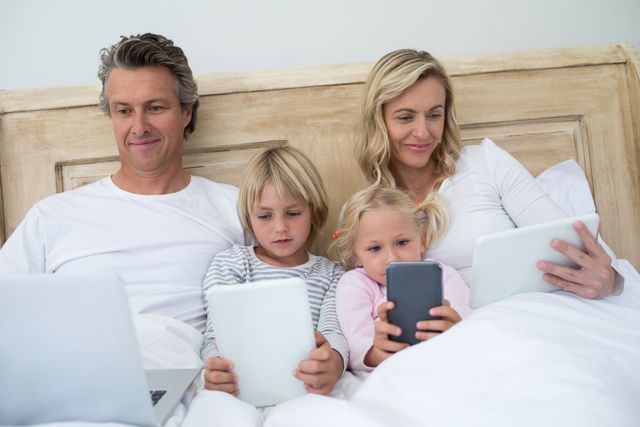 Family of four relaxing in bed, each using a different digital device. Parents and children are engaged with a laptop, tablet, and smartphone. Ideal for illustrating modern family life, technology use, and digital bonding moments.
