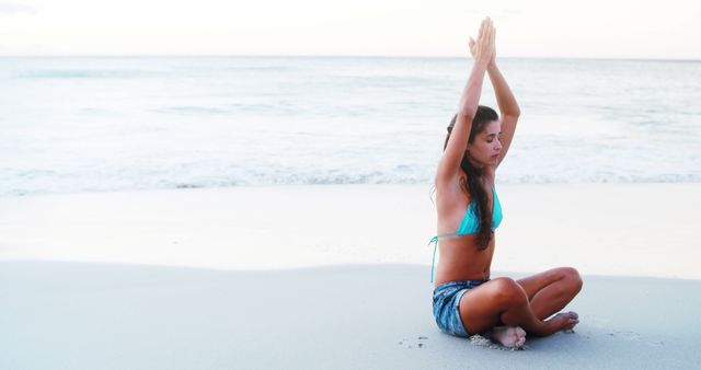 This image depicts a young woman sitting cross-legged on the beach, meditating during sunrise. She is wearing a bikini and yoga shorts, her hands raised above her head in a yoga pose. The calm ocean and serene background create a peaceful atmosphere. Perfect for use in wellness blogs, meditation applications, fitness advertisements, or travel brochures promoting relaxation and natural beauty.