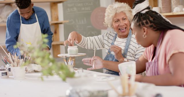 Senior woman teaching younger women pottery techniques in a vibrant art workshop setting. They are engaging with various pottery tools and paints, sharing creative ideas, and building skills. Perfect for articles and promotional materials focused on artistic expression, community engagement, intergenerational activities, and creative hobbies.