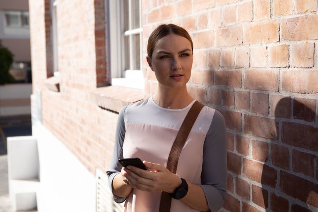 Caucasian businesswoman standing by a brick wall in a city street, holding a smartphone. Ideal for use in business, technology, and urban lifestyle contexts. Perfect for illustrating modern communication, professional women, and on-the-go business scenarios.