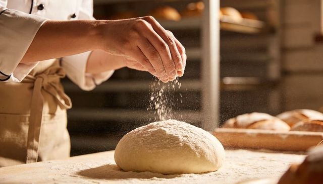 Artisan baker dusting flour over round bread dough on wooden table in warm bakery