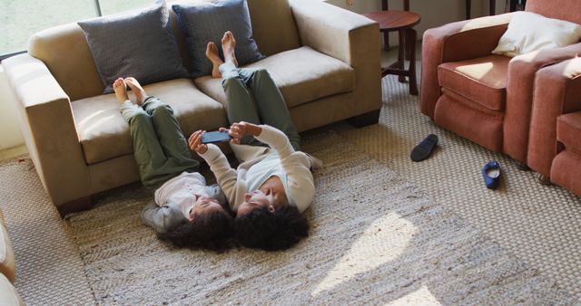 This heartwarming scene captures a mother and daughter lying on the living room floor, engaging with a smartphone. Perfect for promoting family bonding, modern home living, casual lifestyle, and using technology for leisure activities.