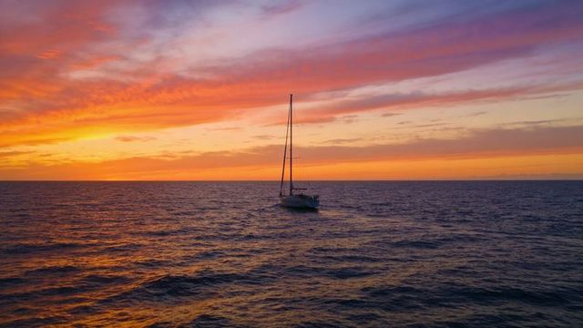 Drifting sailboat at colorful sunset over calm ocean, lone yacht silhouetted on horizon