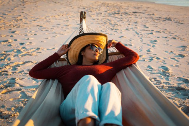 Woman lying in hammock on sandy beach during sunset, wearing sunglasses and hat. Ideal for travel blogs, vacation advertisements, leisure and relaxation themes, and summer promotions.