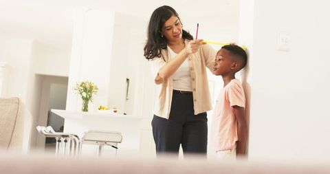 Mother measuring son's height against wall using tape measure in bright open-plan kitchen
