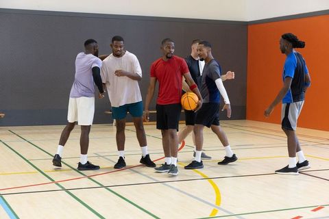 Diverse male teammates gathering on indoor basketball court holding ball and chatting