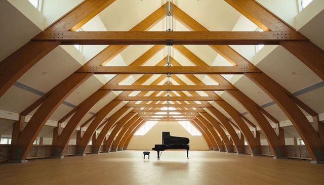 Black grand piano centered in vaulted timber hall with skylights and symmetrical arches