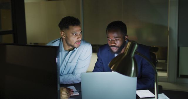 Shows two businessmen working late night in office with computers and desk lamp. Ideal for use in corporate presentations, blog posts about teamwork or productivity, articles on modern work culture, website banners highlighting work ethics or late hours in corporate careers.