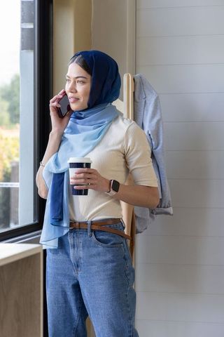 Young woman wearing blue ombre hijab standing by window holding coffee and smartphone with smartwatc