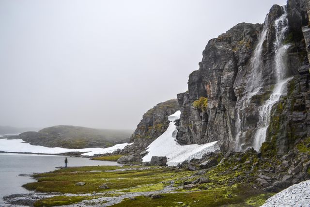 Serene scene featuring a lone fisherman standing near a snow-covered cliff with a cascading waterfall in a misty landscape. Background includes rolling hills and patches of snow, conveying a sense of solitude and peace. Ideal for themes of outdoor adventure, nature's tranquility, isolation, and the beauty of wild environments.