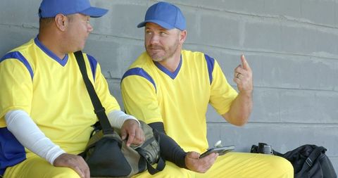Baseball teammates chatting on dugout bench in bright yellow uniforms holding smartphone