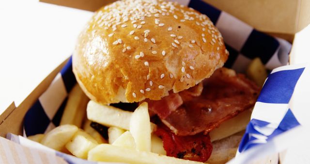 A delicious burger with sesame seeds on the bun, accompanied by crispy bacon and golden fries, is presented in a takeout box. Ideal for showcasing a quick and tasty meal option, this image captures the appeal of fast food.