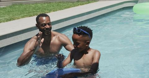 African american father teaching son swimming and playing with kickboard in backyard pool