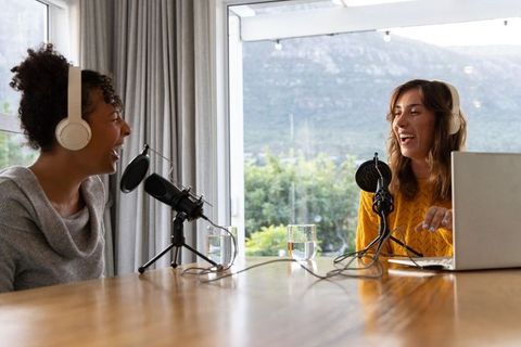 Diverse female co-hosts recording lively podcast at home studio with headphones and laptop