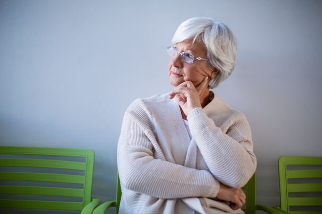 Senior woman with white hair and glasses sitting on a green chair in a hospital waiting room, appearing thoughtful and contemplative. Useful for themes related to healthcare, aging, patient care, medical facilities, and senior wellness.