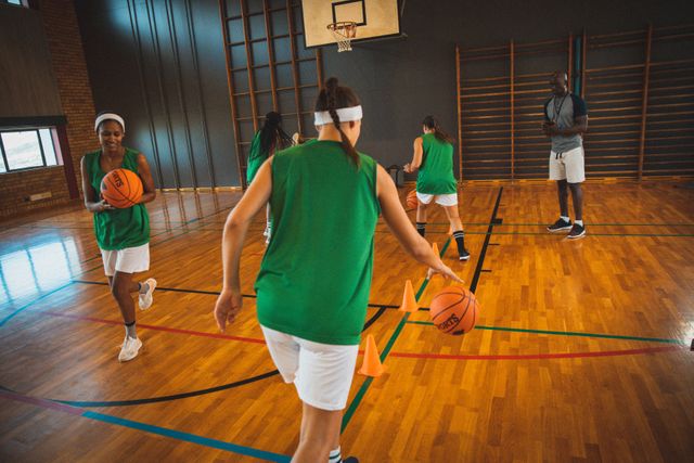 Diverse female basketball team practicing dribbling skills in an indoor court, with a male coach observing their progress. Ideal for use in articles or advertisements related to sports training, teamwork, women's athletics, and physical fitness programs.