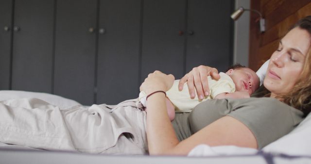 Mother lying down in bed holding her newborn baby who is resting on her chest. This could be used in articles or advertisements focused on motherhood, parenting, family life, or relaxation. Scenes of bonding moments can also be suitable for healthcare and wellness content.