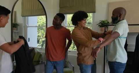 Tailor measuring chest for custom suit while friends helping in sunlit living room