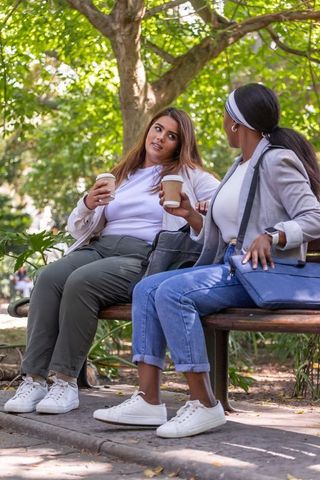 Friends chatting on sunlit park bench holding takeaway coffee cups, casual outdoor conversation