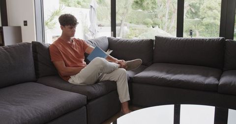 Young man reading paperback on modern sectional sofa in bright minimal living room