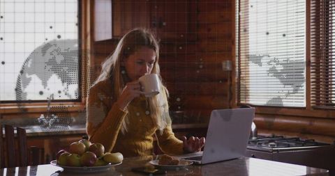 Woman sipping coffee while working remotely in cozy rustic cabin kitchen with laptop