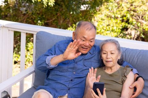 Senior asian couple waving during smartphone video call on sunny porch, enjoying retirement