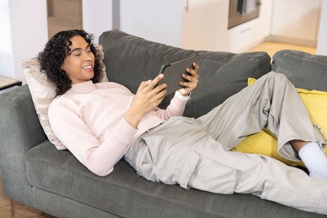Non-binary person reclining on gray sofa using tablet at home for cozy digital relaxation