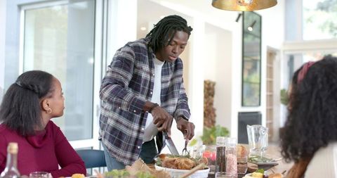 Young Black man carving roast at cozy family dinner in modern sunlit open-plan home