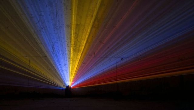 Hooded silhouette facing radiating neon beams in concrete tunnel with cinematic light rays