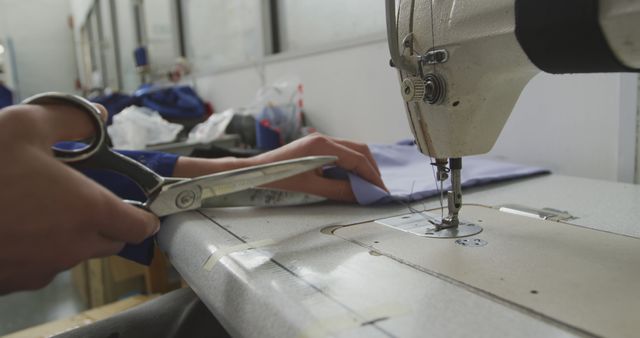 Hands tailoring garment using industrial sewing machine and scissors in clothing production factory. Ideal for content about fashion industry, garment manufacturing, craftsmanship, or industrial sewing.