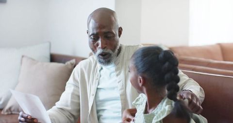 African american grandfather reading letter with granddaughter on cozy living room sofa
