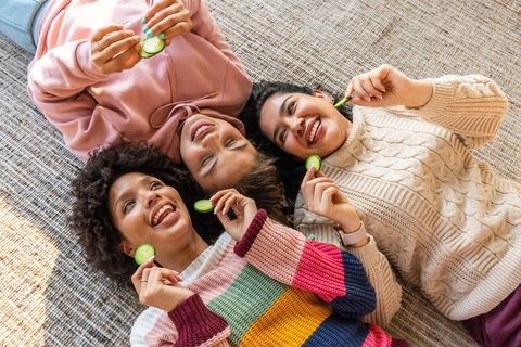 Three friends laughing and relaxing on rug holding cucumber slices for cozy self-care