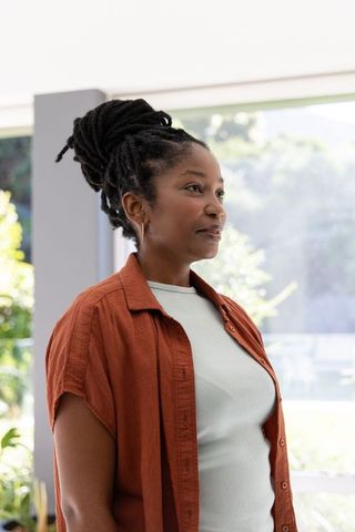 African woman standing by window wearing rust-orange shirt reflecting in sunlit modern home