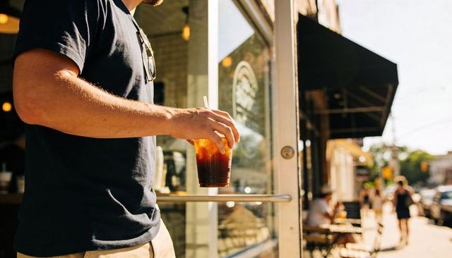 Man standing at cafe door holding iced coffee, summer urban lifestyle and sidewalk vibes