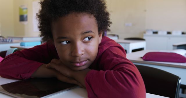 Bored african american schoolgirl leaning on desk looking to window in elementary school classroom. Childhood, education, learning and elementary school, unaltered.