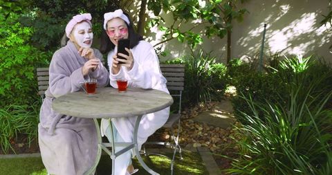 Mother and Daughter in Facial Masks Enjoying Outdoor Time with Tea and Smartphone