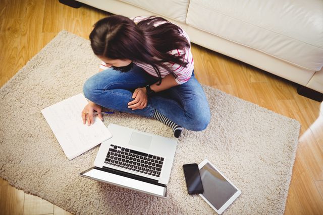 Young woman sitting on carpet with laptop, notebook, tablet, and smartphone, engaging in study or remote work. Ideal for illustrating concepts of online education, remote work, technology use, and modern lifestyle.