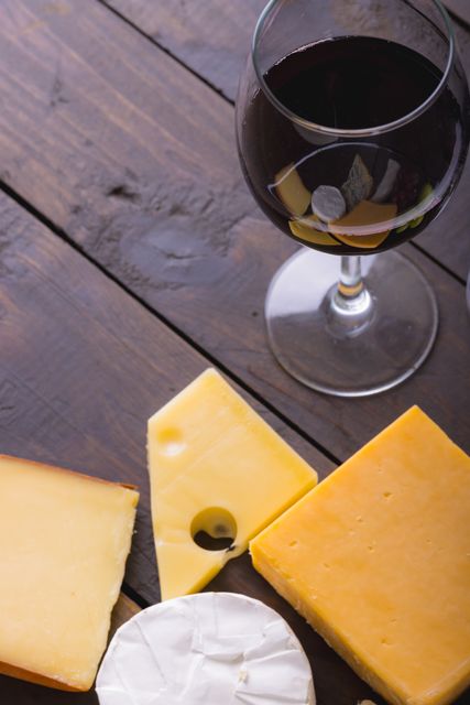 High angle view of various cheeses and a glass of red wine on a wooden table. Ideal for use in food and drink blogs, gourmet recipe websites, restaurant menus, or advertisements for wine and cheese pairings. The rustic setting enhances the appeal for culinary and lifestyle content.
