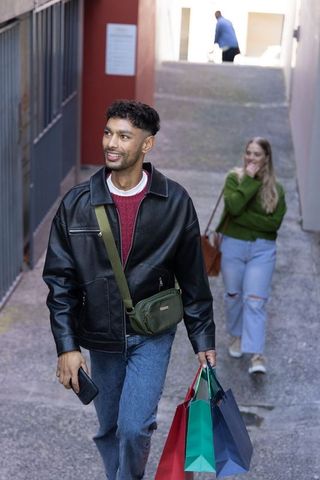 Young indian man walking up urban alley carrying colorful shopping bags using smartphone