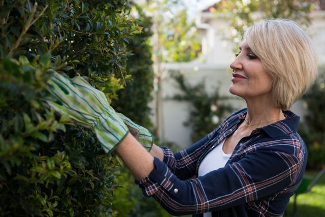 Woman pruning plants in a garden on a sunny day, wearing gloves and a casual outfit. Ideal for use in articles or advertisements related to gardening, outdoor activities, hobbies, and lifestyle. Can be used to promote gardening tools, outdoor wear, or home and garden products.
