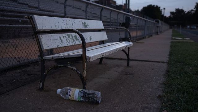 Worn white metal bench with peeling paint and discarded bottle on sidewalk at dusk