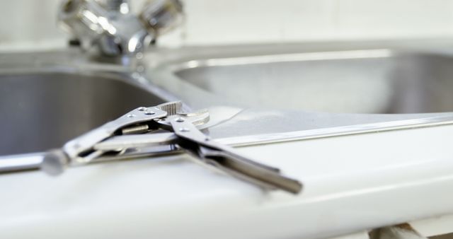 This image shows adjustable pliers resting on the edge of a kitchen sink, suggesting a scene of plumbing repair or maintenance work. The stainless steel sink and faucet are slightly out of focus in the background, highlighting the pliers as the main subject. This image can be used for content related to home improvement, DIY projects, plumbing services, or repair tutorials.