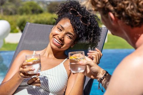 Couple clinking citrus water glasses poolside, woman smiling, lounging in striped swimsuit