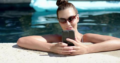 Young woman relaxing on pool edge checking smartphone wearing sunglasses and swimwear