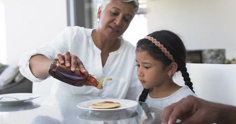 Caregiver pouring maple syrup onto pancake while child watching at sunlit kitchen