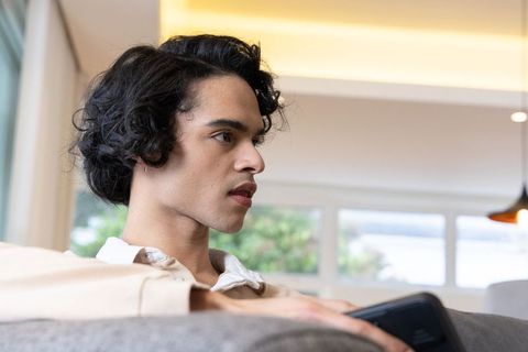 Young man relaxing on gray sofa holding smartphone in modern bright living room side profile