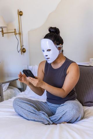 Woman wearing led facial mask while scrolling smartphone on bed for at-home skincare